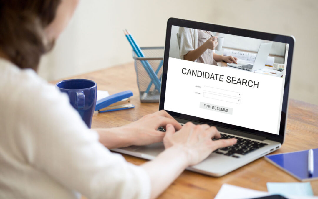 A woman sitting at a desk and looking at a laptop screen that reads, "Candidate Search."