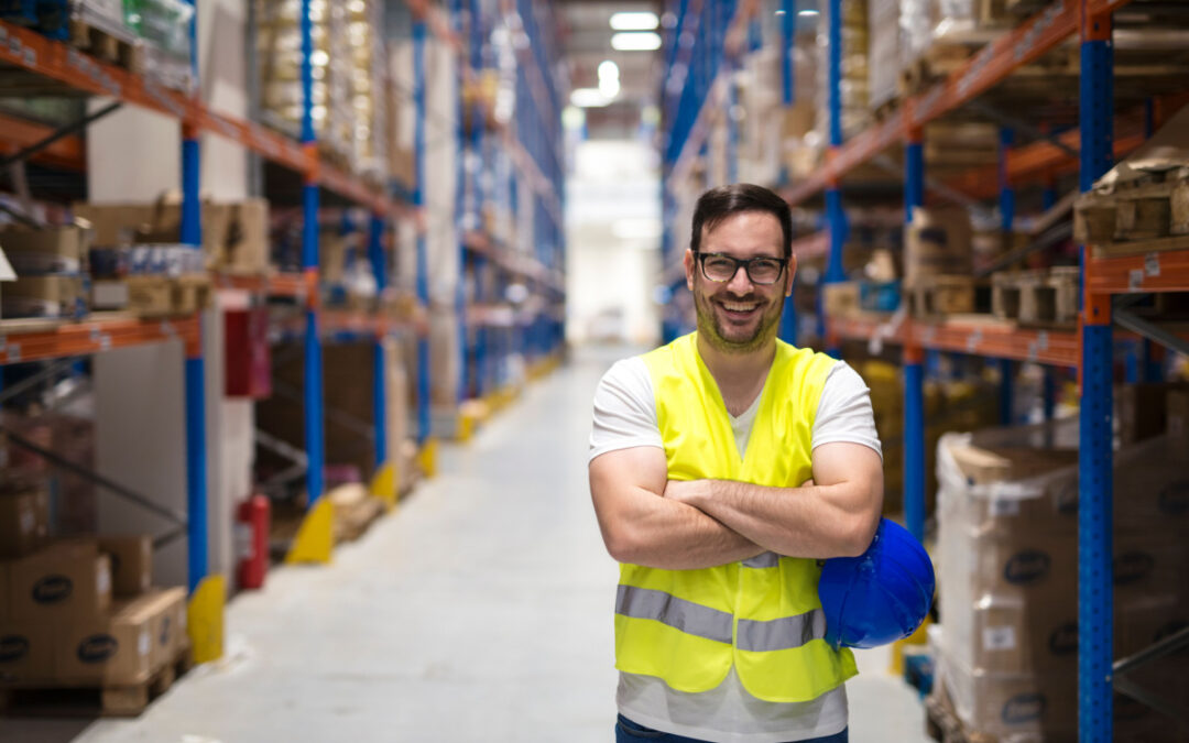 A man in glasses and a high-visibility vest crossing his arms and smiling in front of a warehouse aisle.